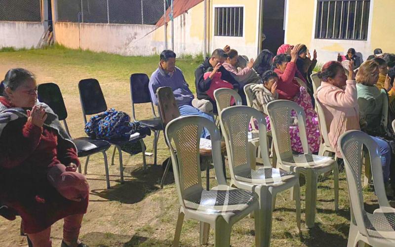 A group of people sit in chairs with eyes closed and hands raised